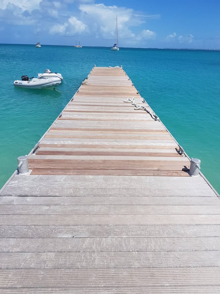 A wooden dock extends over clear turquoise waters, leading toward the horizon. Several boats are anchored in the distance, and the sky is bright with scattered clouds, enhancing the tranquil coastal atmosphere.