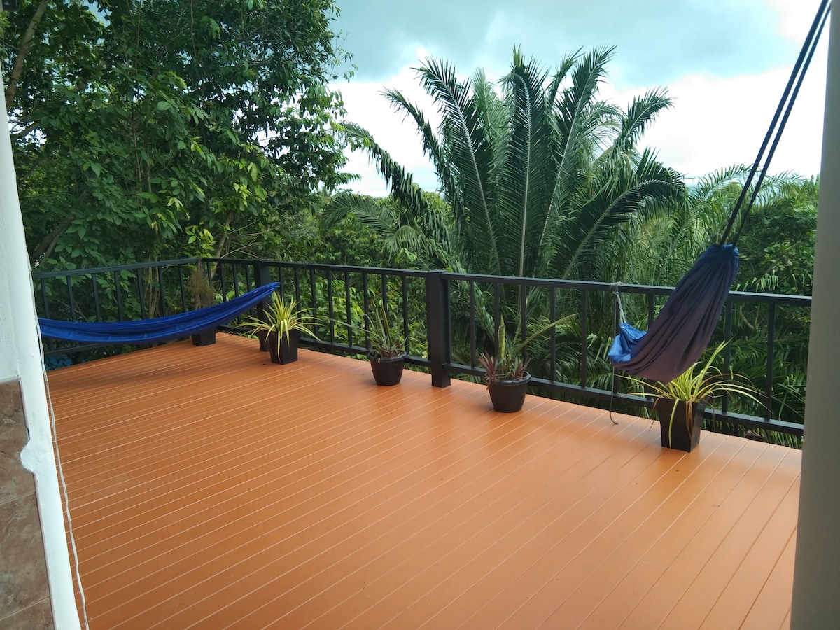 A spacious outdoor terrace features wooden flooring and two hammocks, inviting relaxation. Potted plants are arranged along the railing, adding greenery. Lush tropical foliage is visible in the background, creating a natural, serene setting.