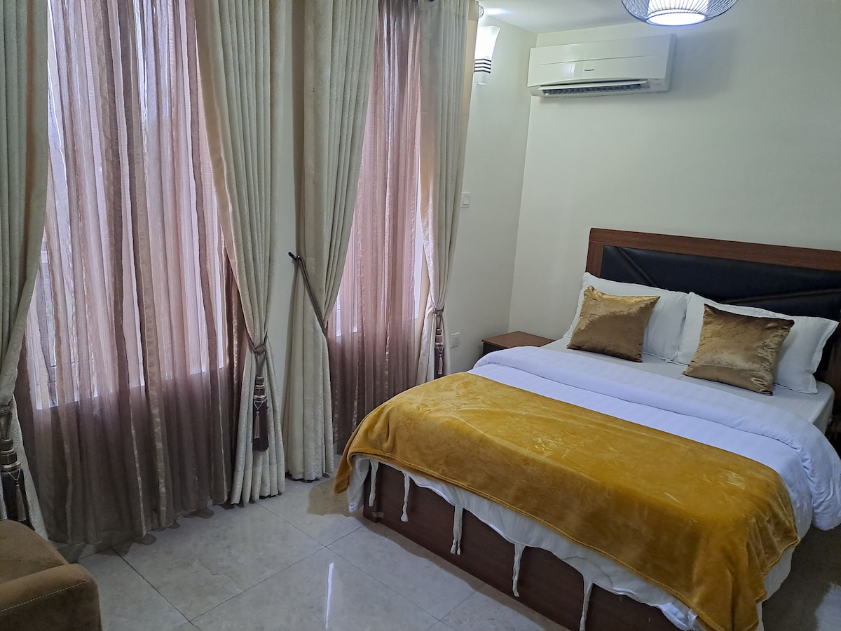 A well-appointed bedroom is presented, featuring a spacious bed covered with a soft yellow throw. Natural light enters through two large, sheer-curtained windows. Neutral tones of the walls and furniture contribute to a calming ambiance, while an air conditioning unit is mounted above the bed.