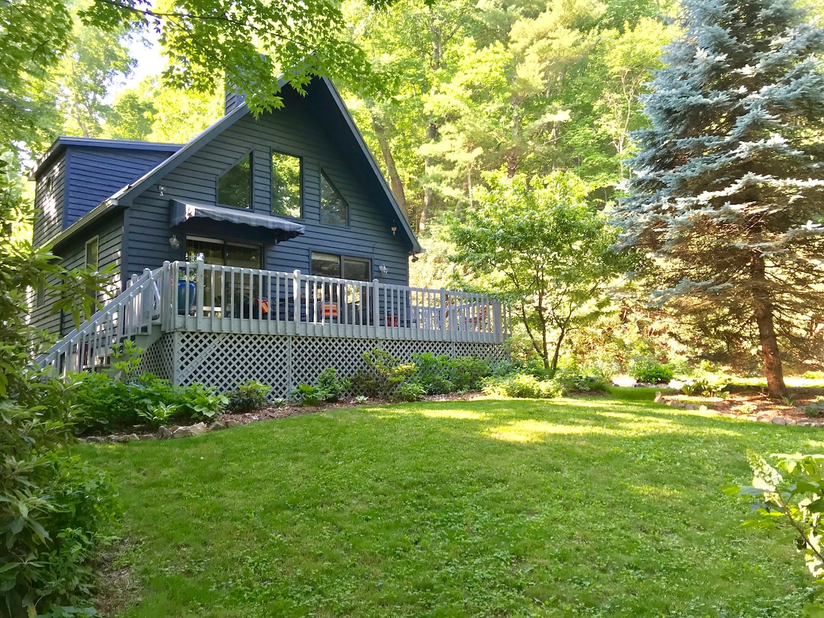 A charming mountain cottage is nestled among lush greenery, featuring a wide wrap-around deck. The deep blue exterior contrasts with the vibrant foliage, while an inviting lawn extends towards the foreground. Towering trees frame the home, enhancing the serene outdoor setting.