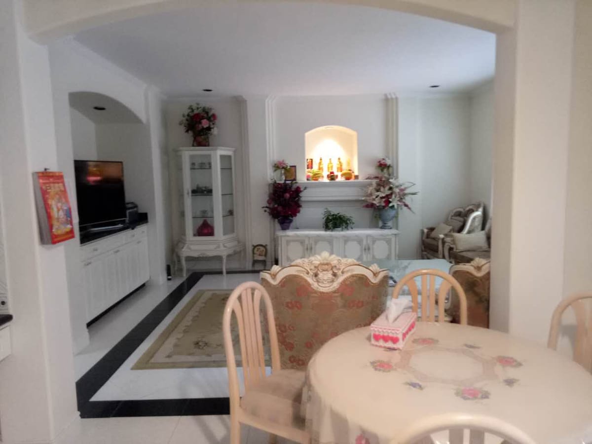 An open-plan living area is visible, featuring a dining table surrounded by four chairs. A comfortable seating area is positioned in the background, with decorative plants and a white cabinet. A television is mounted on a wall, and a fireplace is framed by floral arrangements.