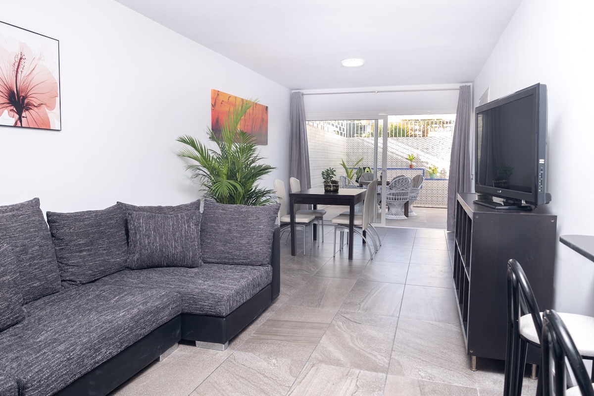 A spacious living area is featured, showcasing a modern sectional sofa and a wooden dining table surrounded by chairs. Large glass doors provide access to an outdoor patio area, while potted plants add greenery to the setting. A flat-screen television is positioned on a black media console.