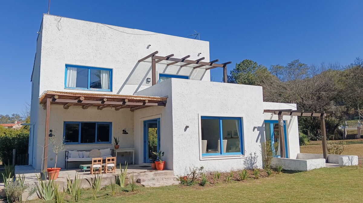 A two-story house is depicted, featuring white walls and blue accents. Large windows invite natural light, while a shaded terrace with outdoor seating enhances the welcoming appearance. Surrounding greenery and a spacious lawn contribute to the tranquil outdoor environment.