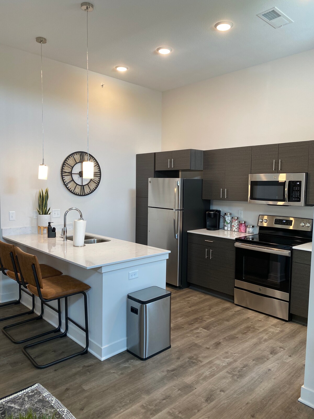 A modern kitchen is equipped with stainless steel appliances, including a refrigerator and microwave. A spacious countertop extends into a bar area with two brown stools. Soft lighting illuminates the space, and a decorative wall clock adds a stylish touch.