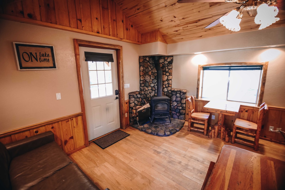 A warm living area features natural wood paneling and a stone fireplace. A small dining space with two wooden chairs is positioned beside a window, while a sofa is set on the opposite side, creating a cozy gathering space. Natural light fills the room.