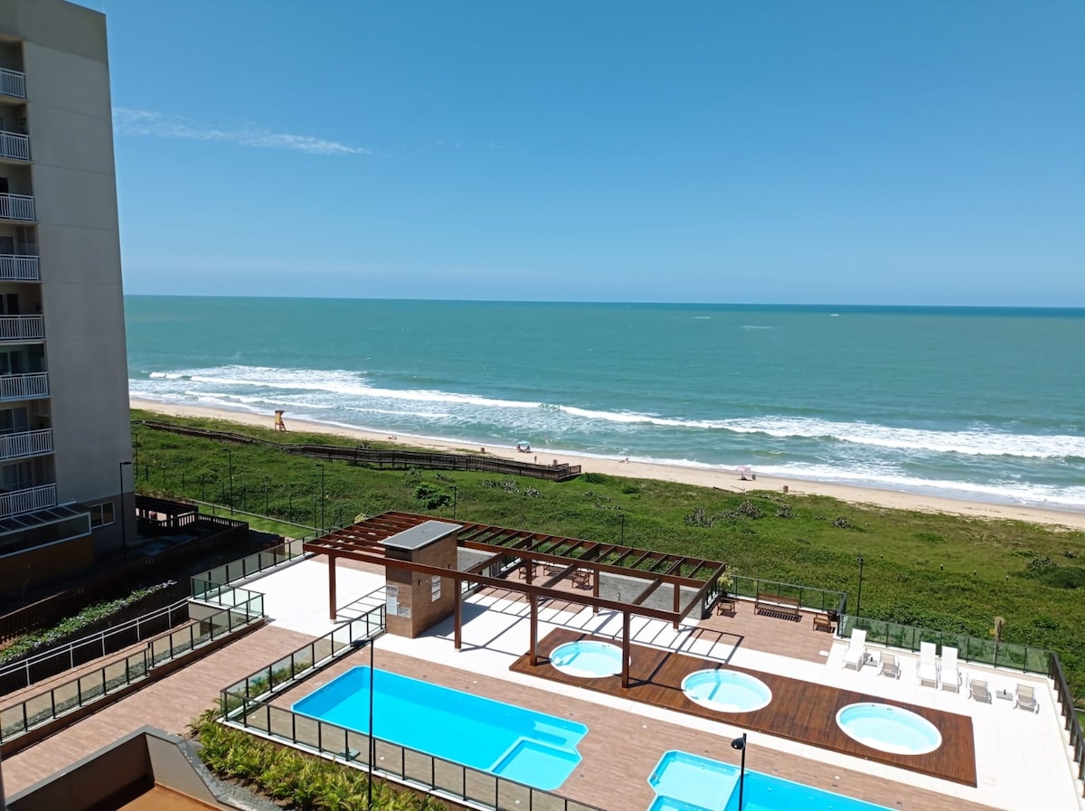 A view of a spacious outdoor area featuring multiple swimming pools, surrounded by wooden decking. A beachfront with gentle waves and sandy shore is visible in the background, complemented by clear blue skies. Lush greenery frames the area, enhancing the serene ambiance.