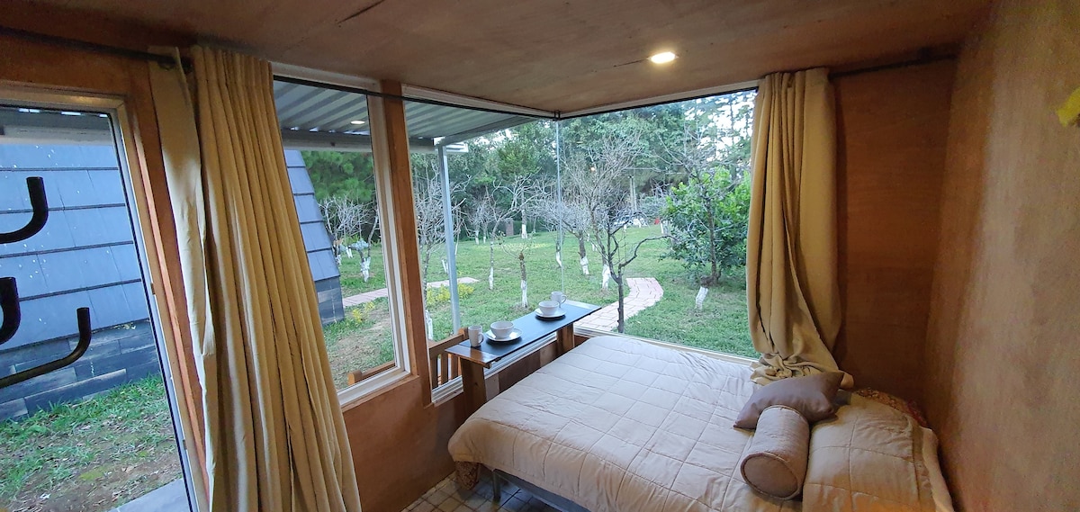 A calming sleeping area features light-colored bedding and a cylindrical pillow. Large windows allow for natural light and views of the greenery outside. A small table is positioned beside the bed, set with two plates, contributing to the inviting atmosphere.