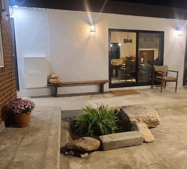 An outdoor space is featured with a small garden area consisting of stones and a plant. A bench is positioned against the wall, providing a resting spot near a window that reveals the interior seating area. Soft lighting accentuates the tranquil environment.
