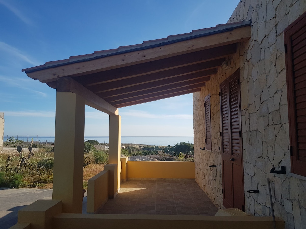 An outdoor patio area is showcased under a wooden roof with beams. The stone exterior of the building is visible, complemented by wooden shutters. A glimpse of the distant sea can be seen against a backdrop of clear blue sky.