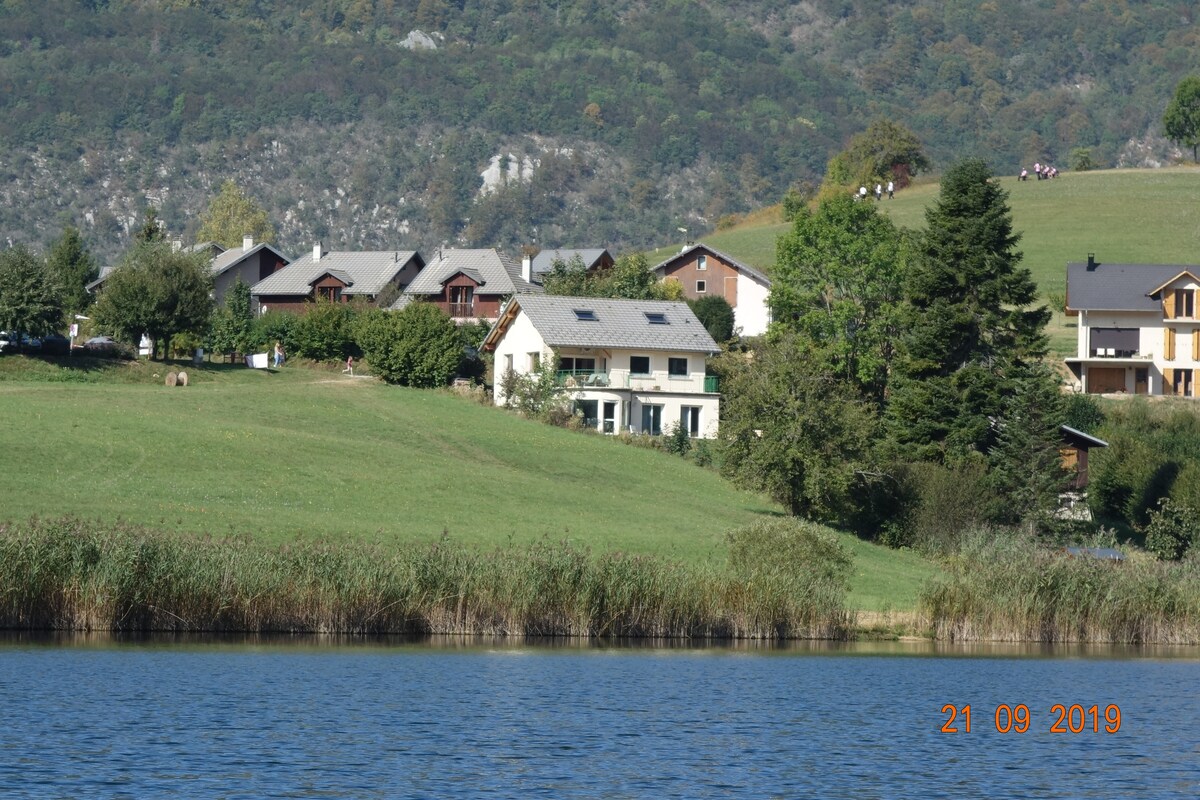 A serene lakeside view captures a cluster of residences set against a backdrop of rolling hills. Lush greenery and a clear sky enhance the natural beauty of the setting, while the calm waters of the lake reflect the surrounding landscape.