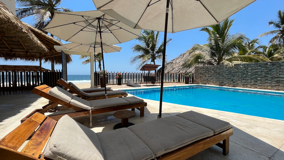 Lounge chairs with soft cushions are positioned around a spacious pool, complemented by large umbrellas providing shade. The clear blue water of the pool reflects the sunny sky, while palm trees are visible in the background, enhancing the tranquil beachfront setting.