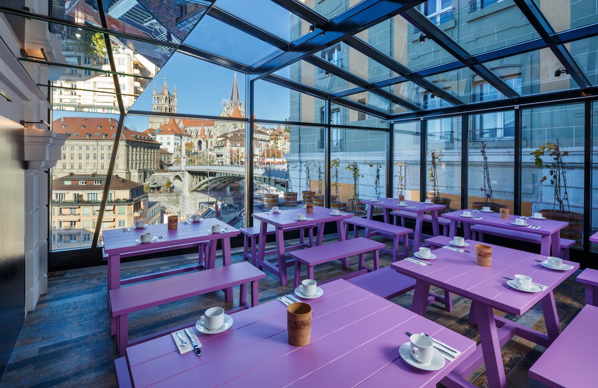 A brightly lit dining area features several purple tables arranged neatly beneath a glass ceiling. The surrounding scenery includes a view of the historic cathedral and cityscape, creating an inviting atmosphere for guests to enjoy their meals.