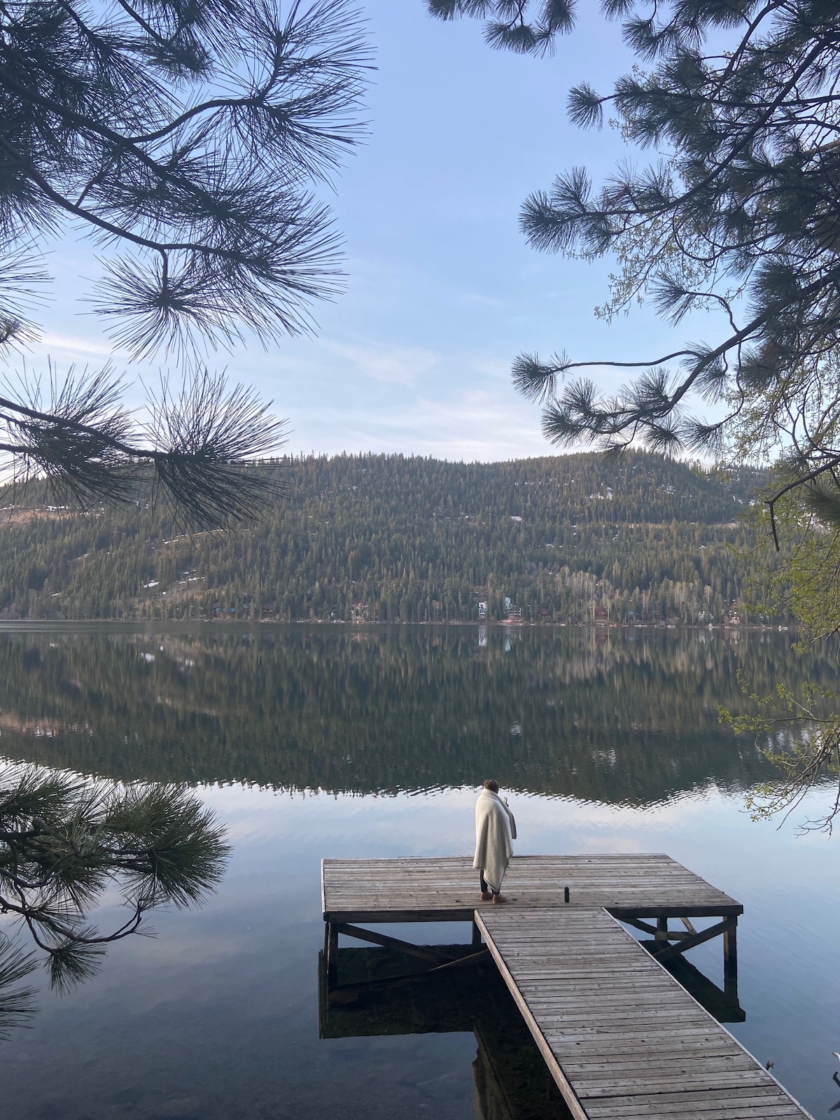 A dock extends into the calm waters of Donner Lake, framed by tall pine trees. Reflections of the surrounding hills and trees can be seen in the still water, creating a serene atmosphere for relaxation.
