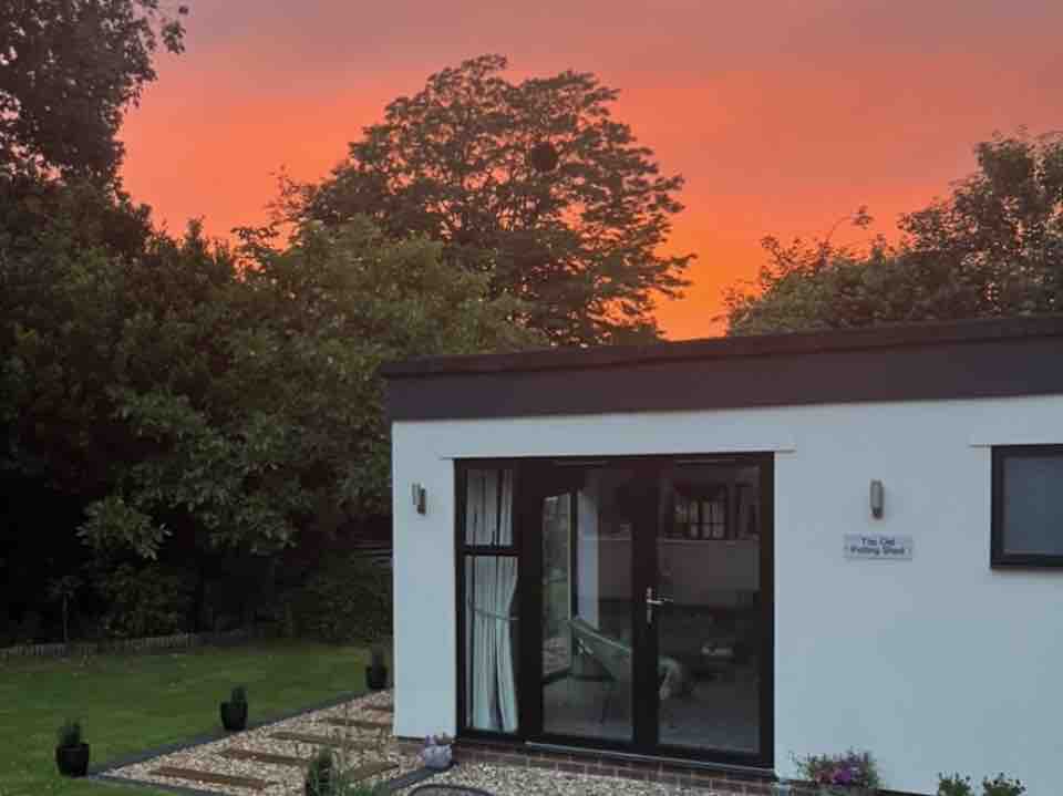 The exterior of The Old Potting Shed is showcased against a vibrant orange sky at sunset. The building features large glass doors, allowing glimpses of the interior. Lush greenery and landscaped gardens surround the space, providing a tranquil atmosphere.