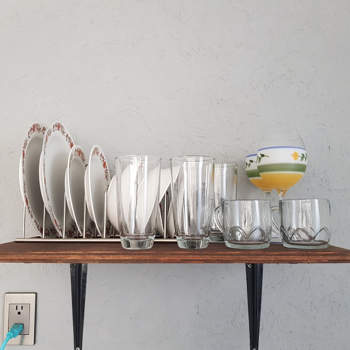 A wall-mounted shelf displays a collection of white plates with floral patterns and various glassware. Six tall glasses stand next to smaller glass cups, with one larger glass featuring yellow liquid and decorative flowers.