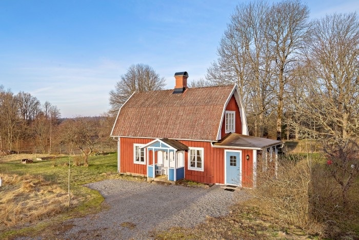 A charming red house with a distinctive sloped roof is depicted. The white-framed doors and windows provide contrast to the vibrant exterior. Surrounding greenery and a gravel path enhance the serene countryside setting, reflecting a tranquil and welcoming environment.
