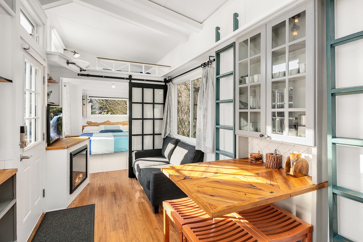 A bright, open living area is featured, showcasing a cozy seating space with a dark sofa and a wooden dining table. Large windows allow natural light to fill the space, revealing a glimpse of the sleeping area with quilted bedding in soft colors.