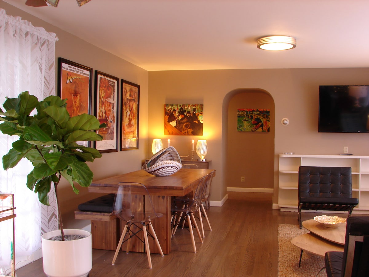 A spacious dining area is highlighted by a large wooden table surrounded by clear chairs. Colorful artwork adorns the walls, and soft lighting adds to the inviting ambiance. A decorative plant in a white pot and a comfortable black chair enhance the overall design.
