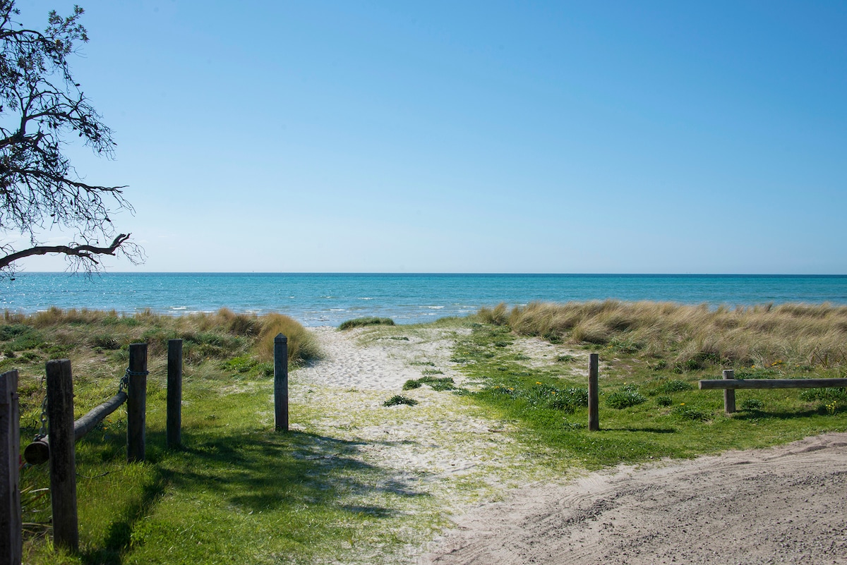 A pathway leads through lush green grass and sandy terrain towards the shimmering blue ocean. Gentle waves can be seen in the distance, framed by soft beach grasses swaying in the breeze. The clear sky adds to the tranquil beach setting.