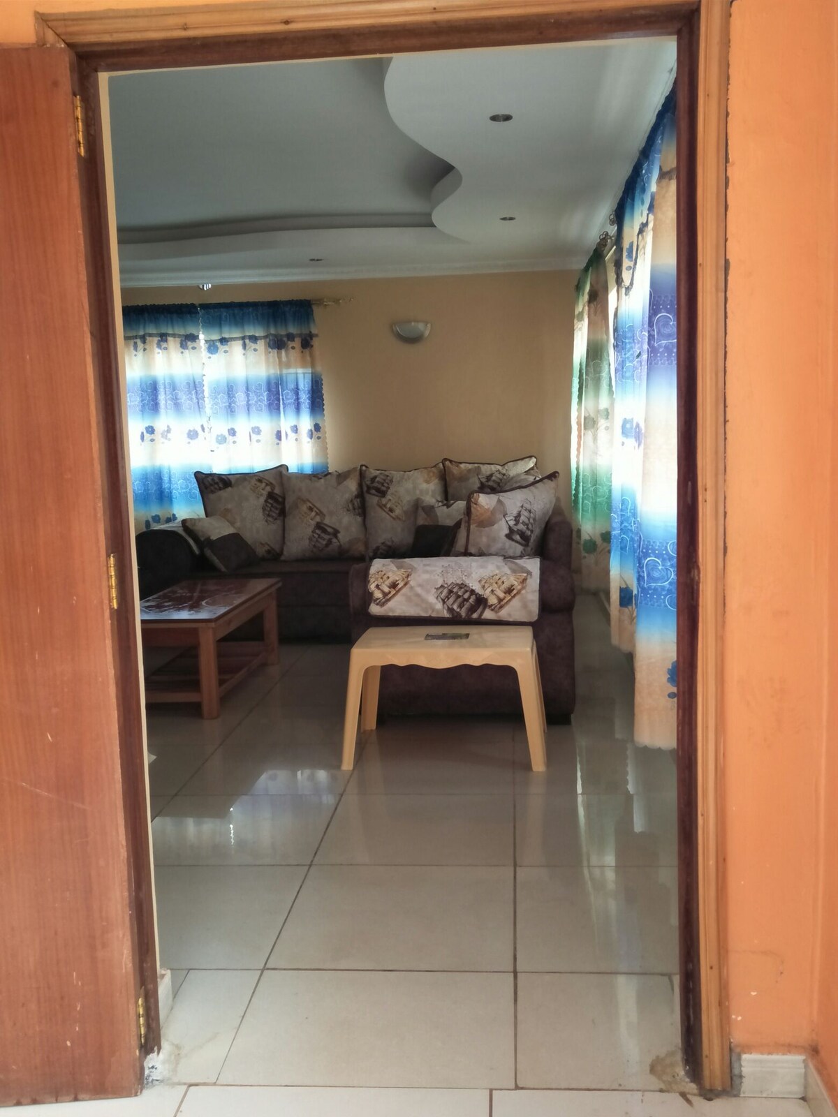 The entrance leads into a living area furnished with a spacious sofa adorned with decorative cushions. A small coffee table is positioned in front of the sofa. Natural light enters through large windows, enhancing the bright ambiance of the room.