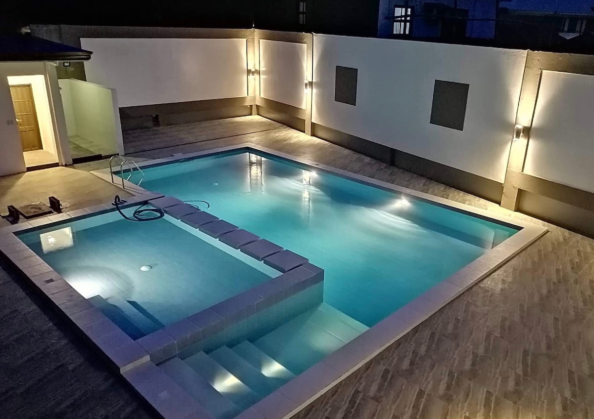 A well-lit swimming pool area features a spacious, rectangular pool with clear water reflecting lights. Surrounding walls are illuminated, creating a calming atmosphere. Steps lead into the pool, and a relaxing seating area is positioned nearby.