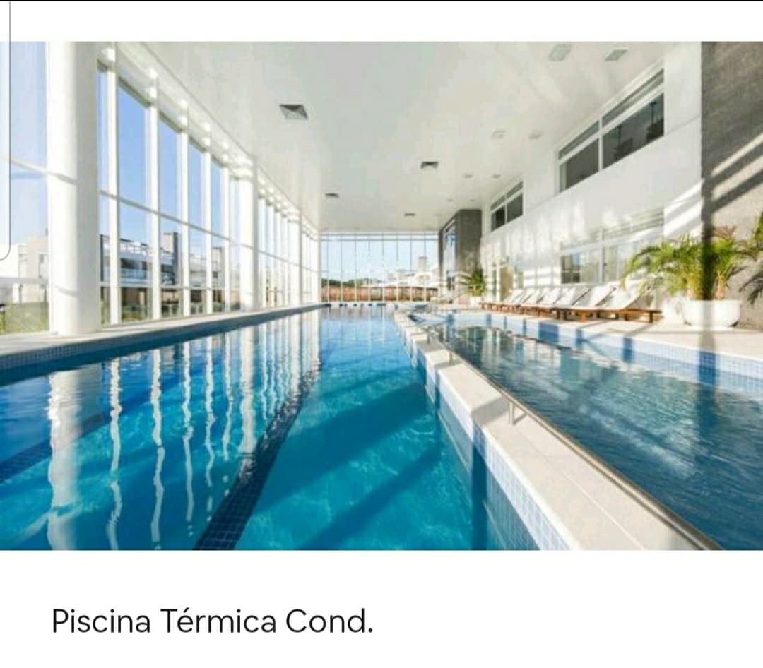 An indoor thermal pool features large windows, allowing natural light to fill the space. The water appears invitingly clear, with lounge chairs lined up along the edges. Potted plants add a touch of greenery, enhancing the serene atmosphere.