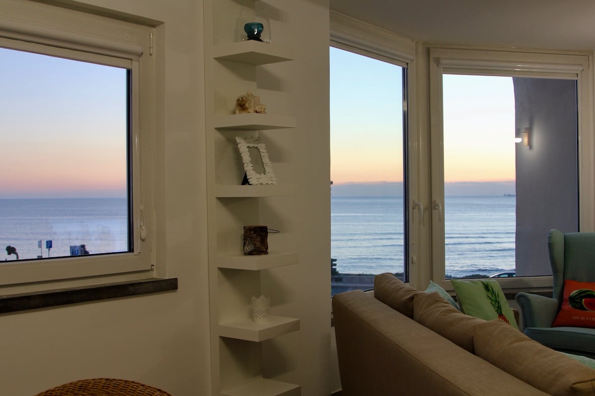 Large windows allow for a sweeping view of the ocean at sunset, where soft hues of orange and pink blend into the sky. A simple white shelving unit is positioned near the window, providing a functional display space for decorative items.