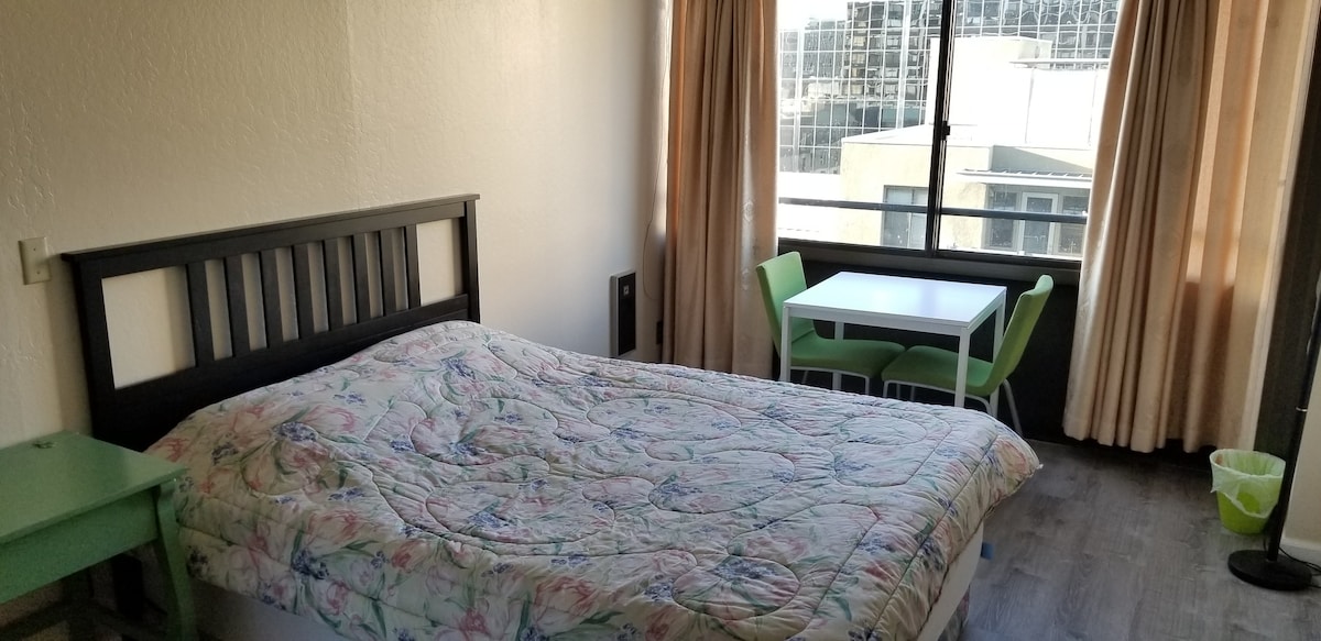 A well-lit room features a spacious bed with a floral quilt and a small dining area with a table and green chairs. Large windows offer views of the outside, while light-colored curtains provide soft illumination.