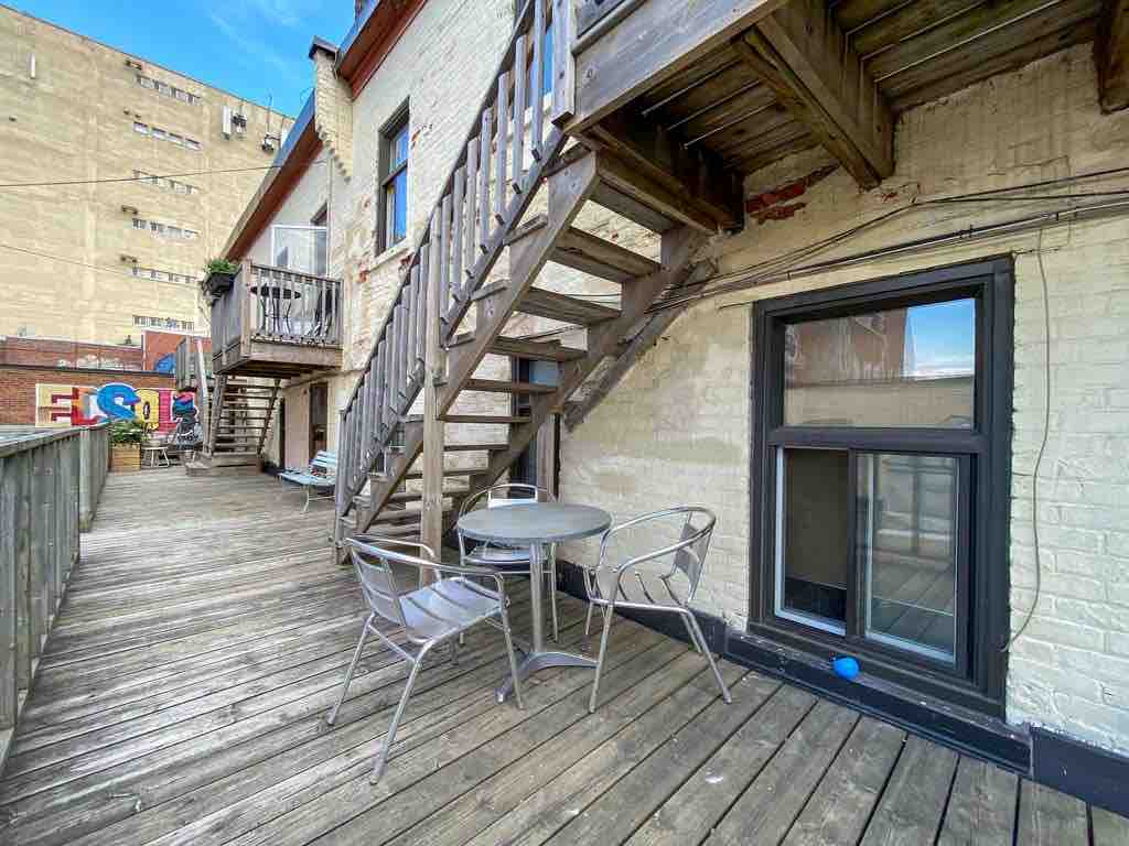 A spacious outdoor terrace is featured, showcasing wooden decking and a small round table with four metal chairs. Staircase access is visible on one side, leading to upper-level units. The surrounding area is lined with a low wooden railing, enhancing the outdoor experience.