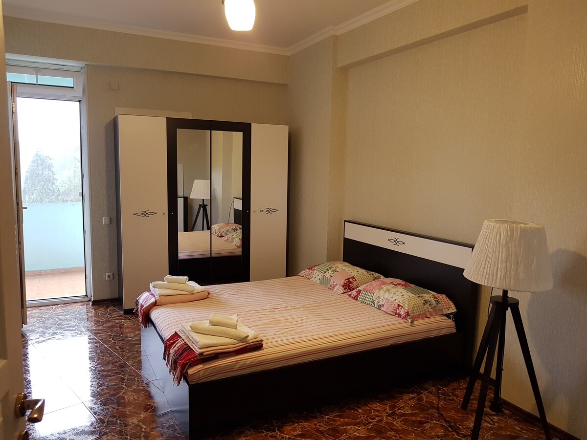 A spacious bedroom features a large bed with decorative pillows and fresh linens. A wardrobe with mirrored doors reflects light into the room. Natural light enters through a sliding glass door that leads to a small balcony. A lamp adds soft illumination.