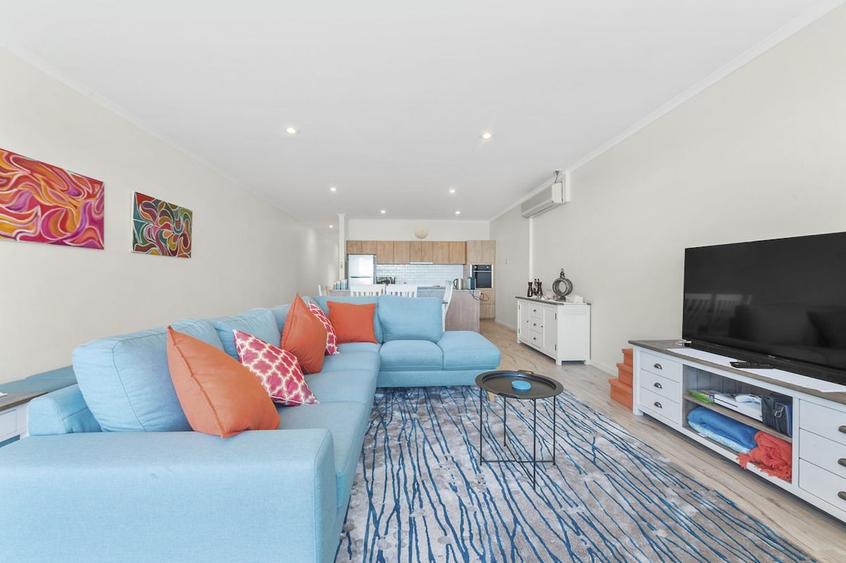 A spacious lounge area is furnished with a large blue chaise lounge adorned with colorful cushions. An LCD TV is positioned on a media console, and artwork adds a pop of color to the light-toned walls. Natural light illuminates the open layout.