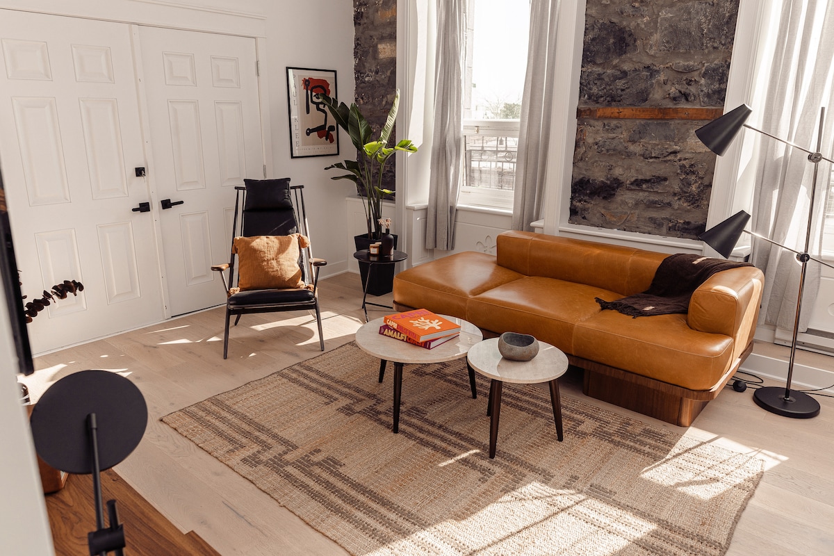The living area showcases a modern leather sofa paired with a minimalist coffee table. A comfortable armchair is positioned next to a leafy plant. Light streams through large windows, highlighting the textured stone wall and wooden flooring, creating a welcoming atmosphere.