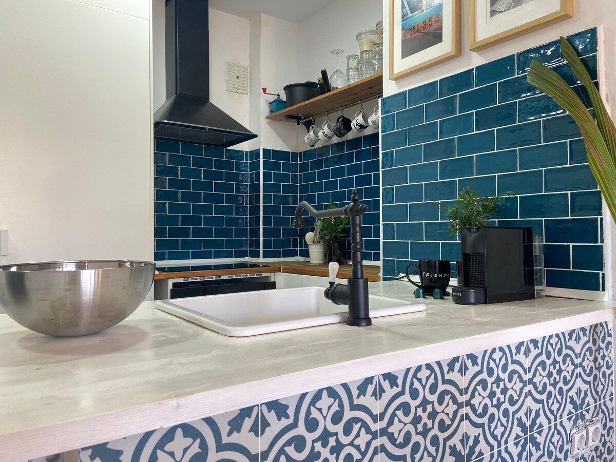 The kitchen area features vibrant blue tile accents on the walls, complemented by wooden countertops. A sleek black faucet and sink are positioned prominently, alongside modern appliances, including a coffee maker. Decorative plants add a touch of greenery to the space.
