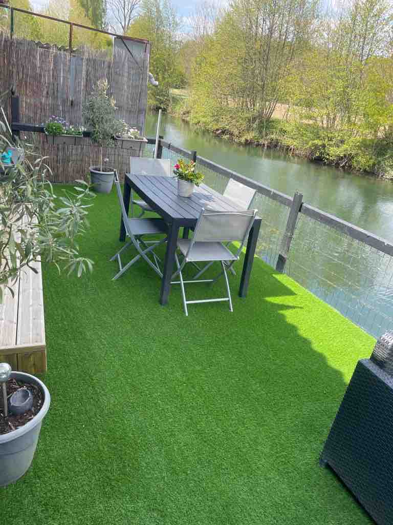 A terrace overlooking the Sèvre features artificial grass flooring and a dining table with chairs. Potted plants are positioned around the area, enhancing the natural vibe. The calm water of the Sèvre can be seen in the background, framed by lush greenery.