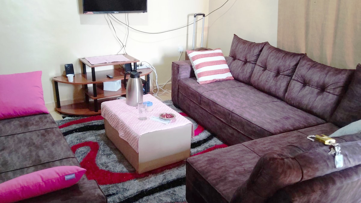 A cozy living area features two sectional sofas adorned with pink accent pillows. A low coffee table sits centrally, accompanied by simple decor items. A mounted television is positioned on the wall, with wires neatly contained, and a warm rug adds texture to the floor.
