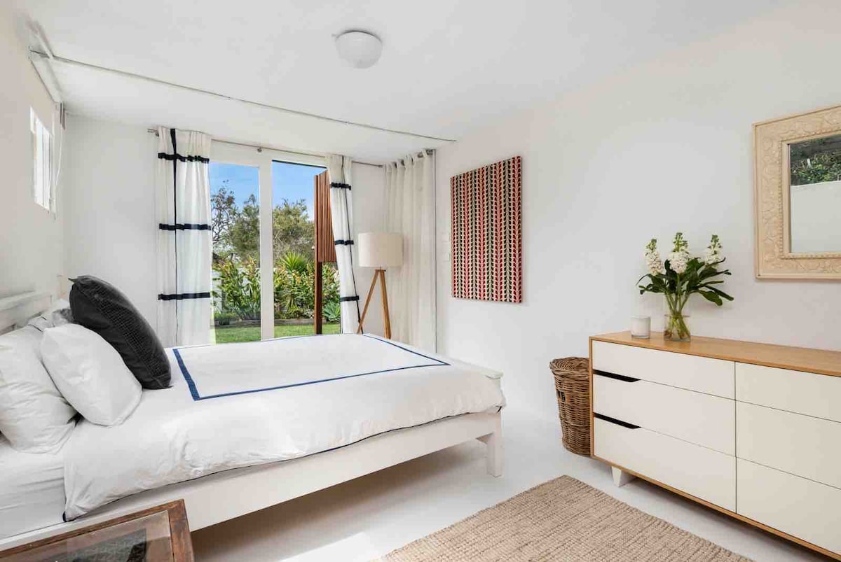 A bright bedroom features a spacious bed with crisp white linens, complemented by patterned pillows. Natural light floods in through large sliding doors, which open to a garden view. A wooden dresser with decorative accents sits nearby, alongside a stylish woven basket and a floor lamp.
