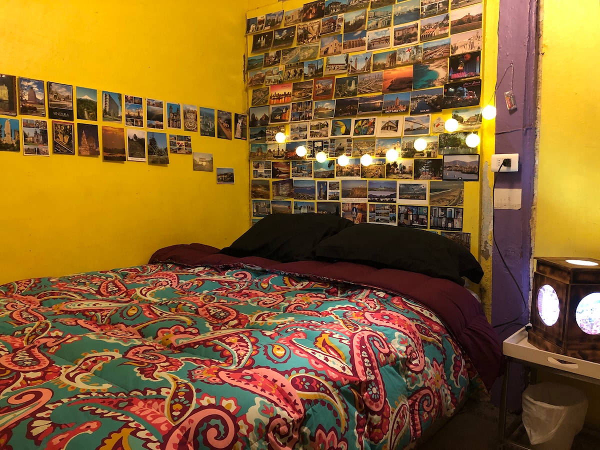A vibrant bedroom is highlighted by a spacious bed covered in an intricately patterned comforter. A collection of postcards adorns the yellow wall behind the bed, with warm lights strung across, creating a cozy ambiance. A bedside table with a unique lamp adds character to the space.