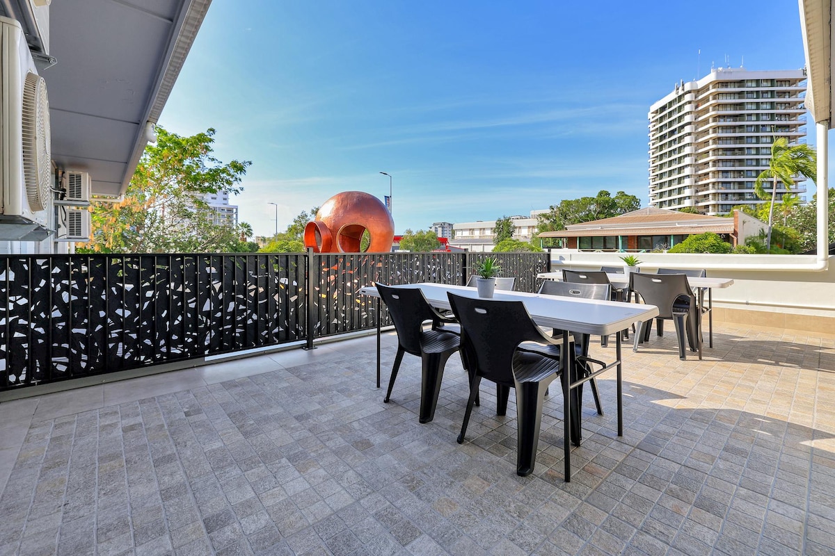 A spacious terrace is presented with outdoor dining tables and chairs. Lush greenery is visible around the area, alongside modern buildings. The terrace features a decorative black railing, providing a contemporary look while the sky above is clear and bright.