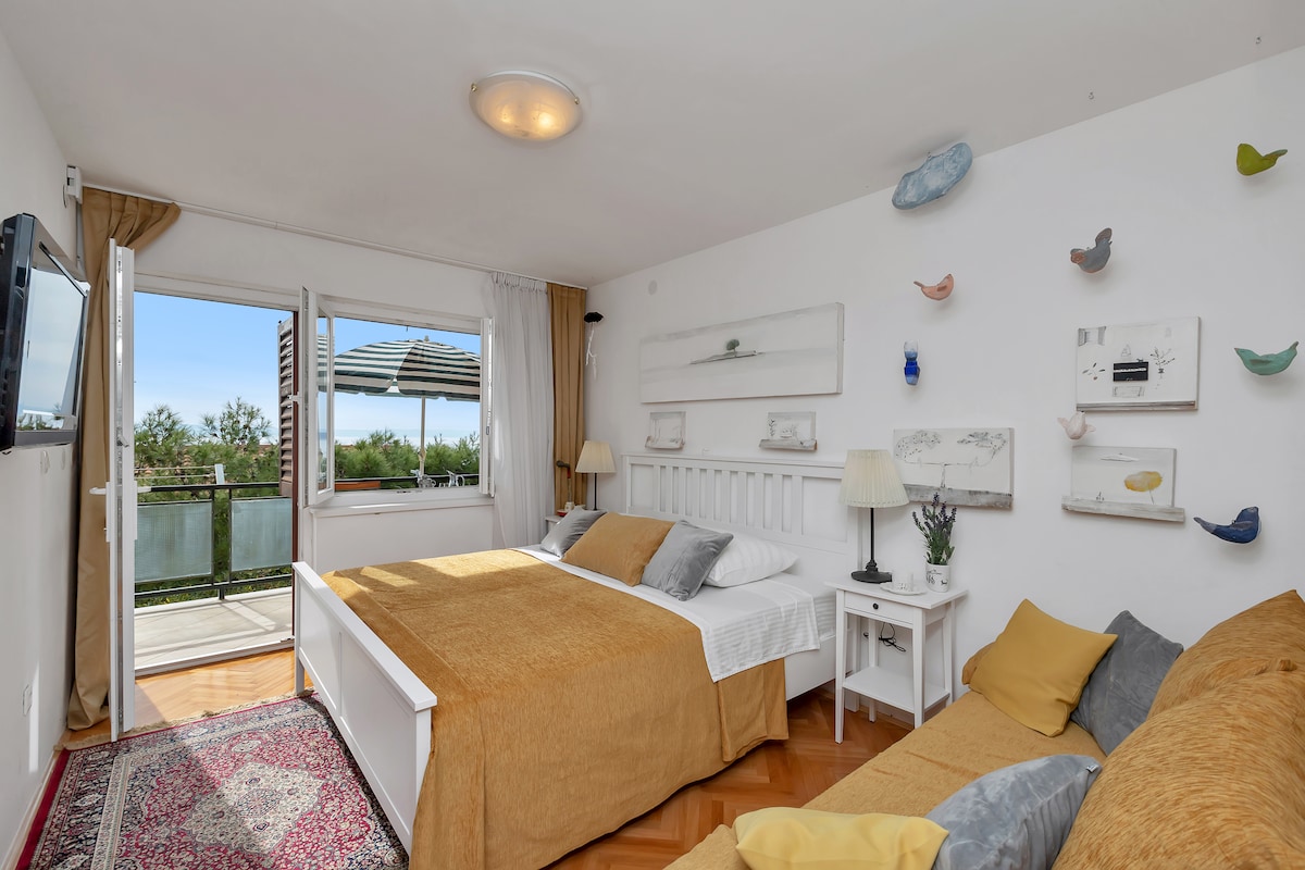 A bright and airy studio apartment is presented with two spacious beds, accented by warm yellow bedding. A comfortable sitting area is visible, complemented by decorative pillows. Large windows provide access to a terrace, with a view of outdoor greenery and a covered seating area.