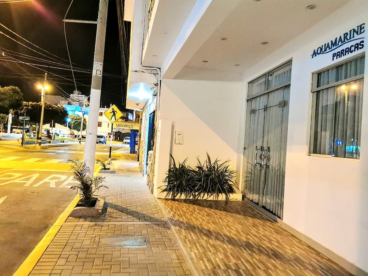 The exterior of the property is visible at night, featuring a well-lit entrance with large glass doors. A neatly paved walkway leads to the entrance, flanked by small plants. Street signs and distant lights from nearby establishments can be seen, indicating a vibrant atmosphere.
