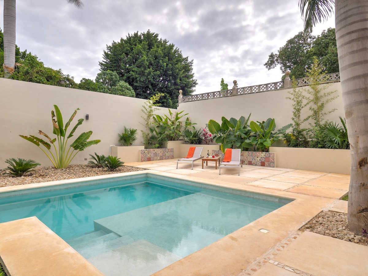 A private pool area is surrounded by lush greenery and decorative landscaping. Two lounge chairs sit by the edge of the pool, inviting relaxation. The pool's clear water reflects the sky, enhancing the tranquil ambiance. Textured stone surfaces provide a clean, contemporary look.