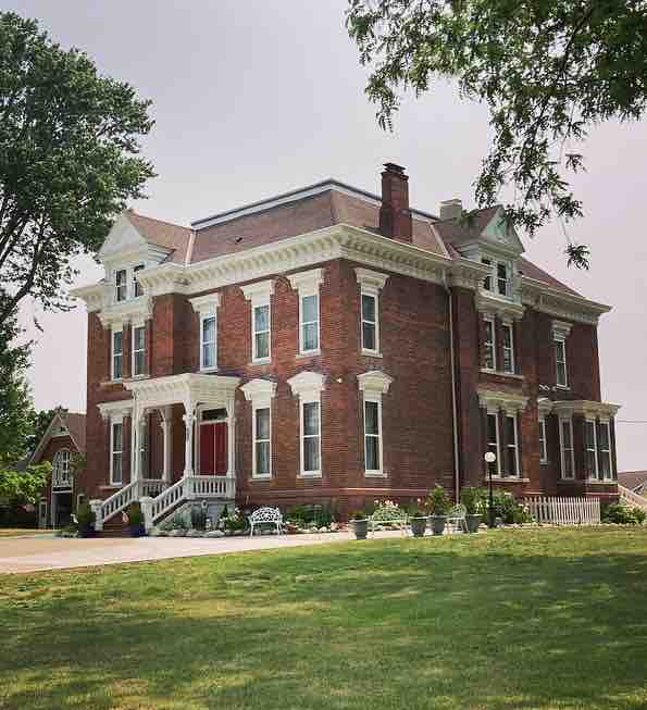 An elegant historical Italianate home, constructed in 1886, features a brick exterior with white trim and intricate architectural details. Tall windows adorned with shutters reflect the period's design. The expansive lawn is framed by trees and flower arrangements, inviting a serene atmosphere.