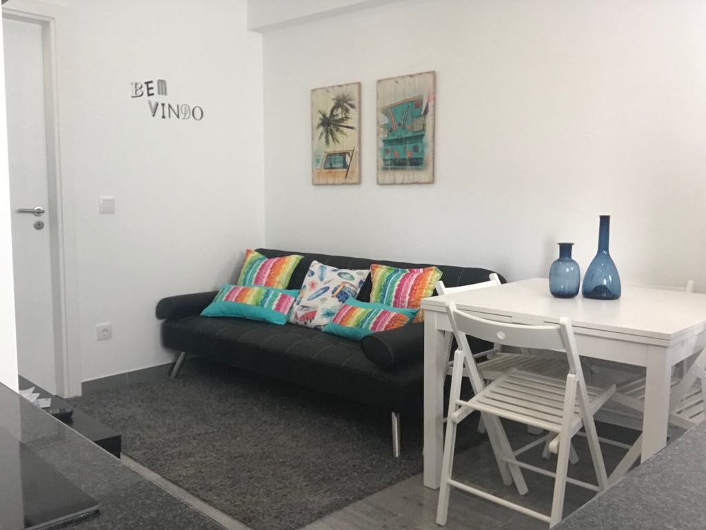 A modern living area displays a sleek black sofa adorned with colorful cushions. A dining table and white chairs are positioned nearby. Light-colored walls enhance the space, while framed artworks add a touch of vibrancy. A welcoming atmosphere is conveyed through thoughtful decor.