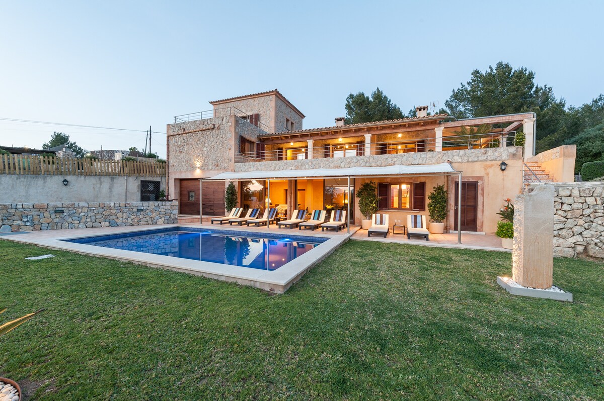 The exterior of the villa showcases a modern design with multiple levels, surrounded by a well-maintained lawn. A saltwater pool is located directly in front, accompanied by comfortable sun loungers and shaded areas. The warm light of dusk enhances the inviting atmosphere of the outdoor space.