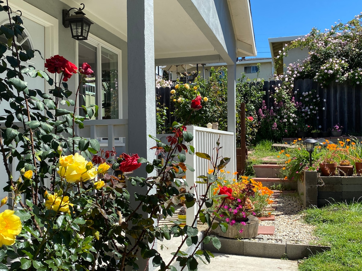 A vibrant garden is highlighted with blooming roses, including yellow and red varieties, framing the entrance of the house. A variety of colorful flowers thrive in the area, with a manicured lawn visible in the background under a clear blue sky.