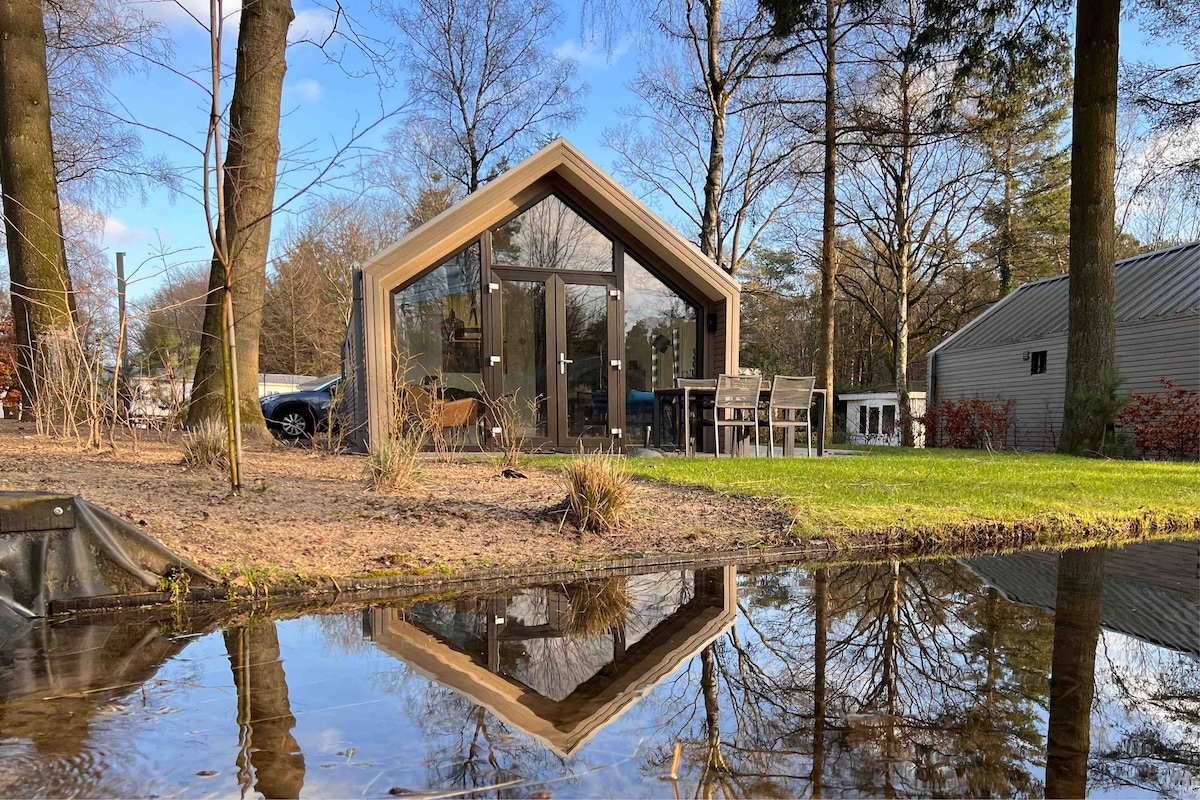 A modern chalet features large glass windows reflecting in a serene pond. Surrounding trees provide a natural setting, with a spacious patio furnished with a table and chairs, inviting outdoor relaxation.