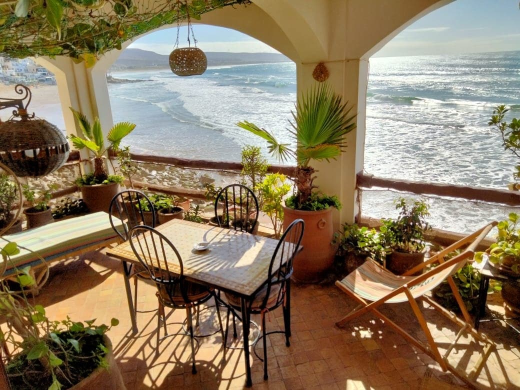 A spacious balcony is adorned with greenery and offers a table surrounded by several black chairs. A hammock rests to one side, while the ocean view features gentle waves lapping at the shore. Sunlight casts a warm glow over the area.
