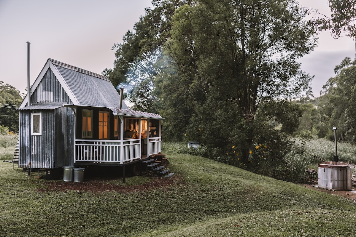 A charming tiny house is set amidst lush greenery, featuring a corrugated metal exterior and a welcoming porch. Warm lights are visible through the windows. Just beyond, a hot tub is situated in the tranquil outdoor space, creating a serene atmosphere in nature.