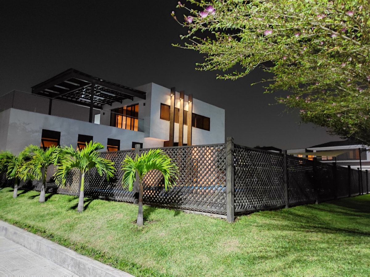 A modern two-story house is surrounded by lush greenery. A well-maintained lawn lies in front, framed by a decorative wooden fence. Large windows reflect ambient light, while a covered patio area is visible on the ground floor, offering a glimpse of outdoor living space.