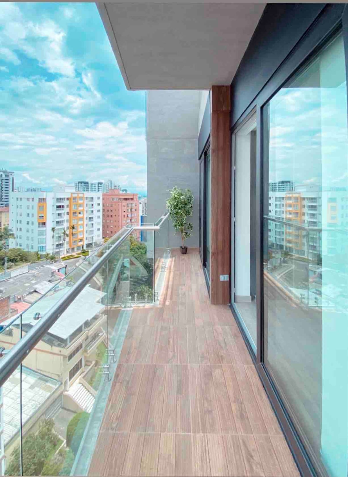A spacious balcony is shown, featuring glass railings and a modern tiled floor. A potted plant is positioned at one end, overlooking a cityscape with various buildings and green spaces in the distance. Clear skies enhance the open feel of the space.